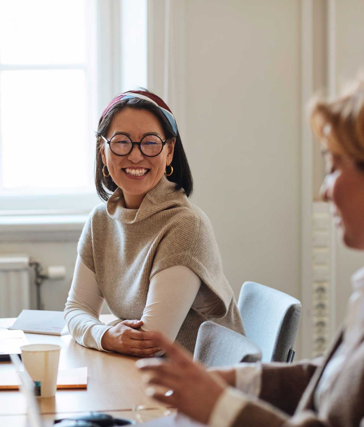 Woman with round black glasses sitting at table smiling while talking to colleague.
