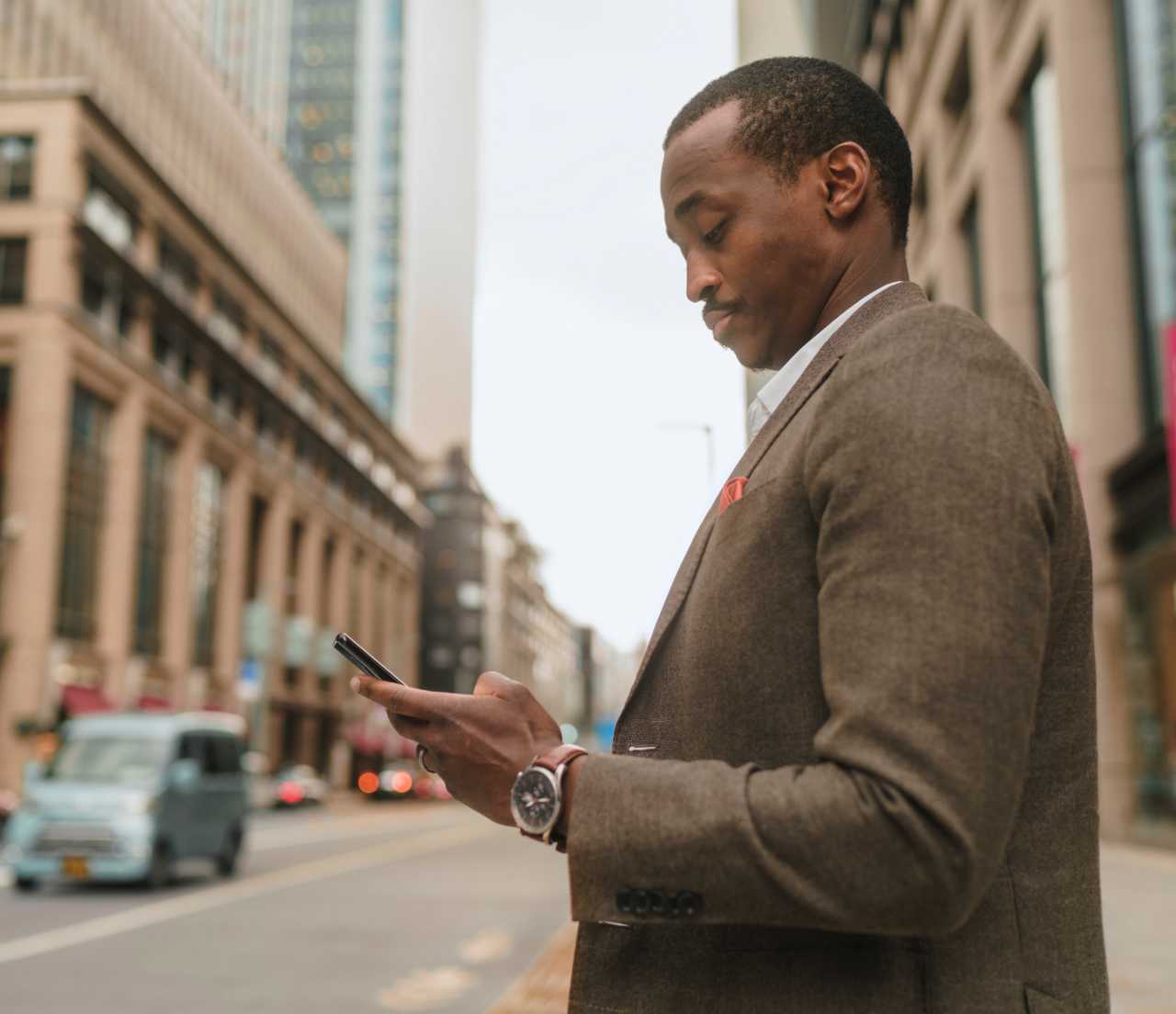Man in suit standing on city street while looking down at smartphone with video play button in center of photo.